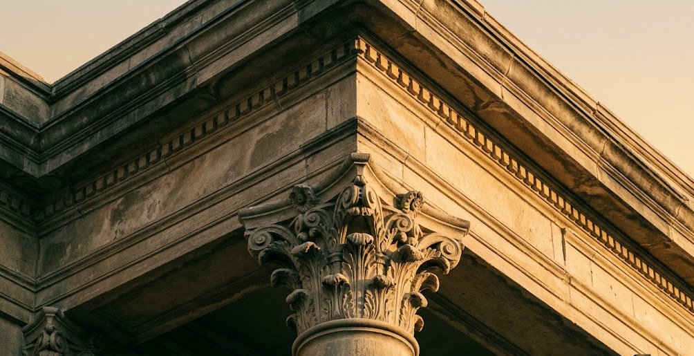 Detail of a weathered limestone Corinthian column capital and entablature in late-afternoon light — carved acanthus leaves, dentil molding, the architecture of institutions built to last.